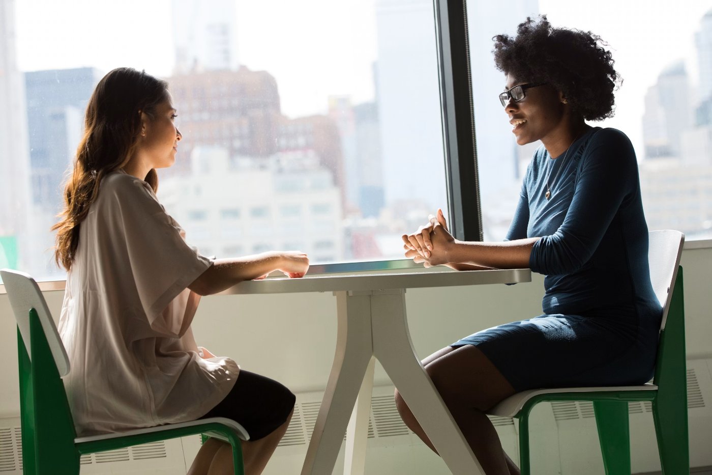 A mentor and a young person in conversation across a table by a window, with the city skyline behind them